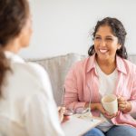 A woman wearing a pink shirt sits on a couch and smiles while holding a cup of coffee. She is talking to another woman sitting next to her, who is wearing a white shirt.